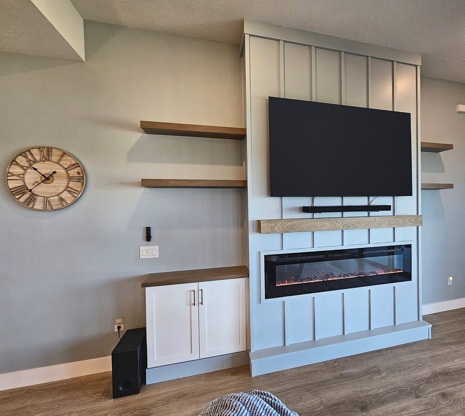Light gray vertical board bump-out media wall with TV, soundbar, wood mantel and linear fireplace, floating wood shelves on both sides and a white base cabinet, in a Venice, FL home